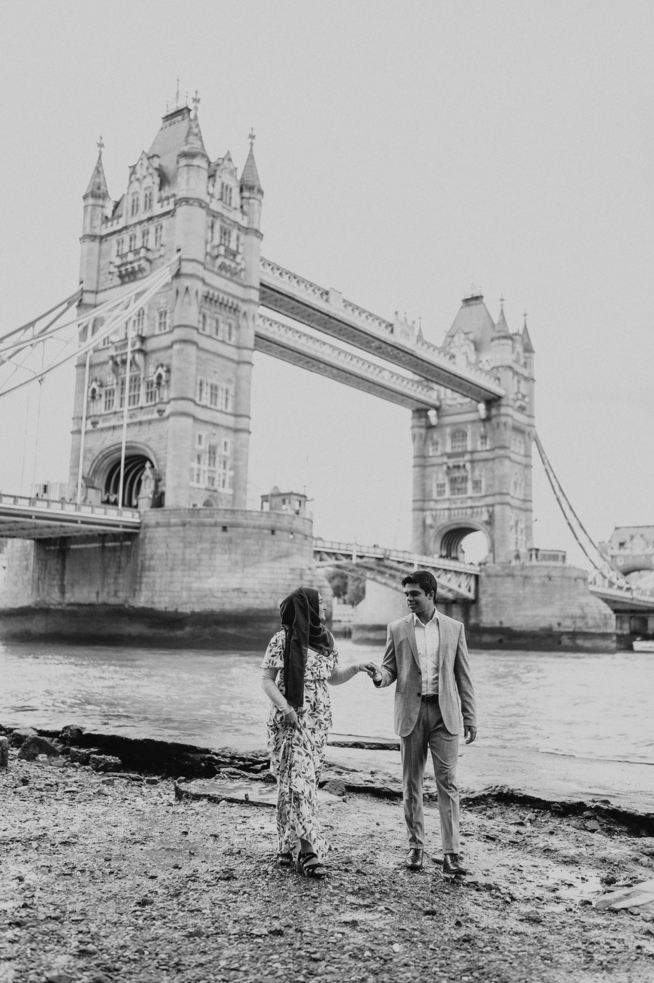 Black-and-white photograph of a couple walking hand in hand beneath Tower Bridge during their London engagement photoshoot.