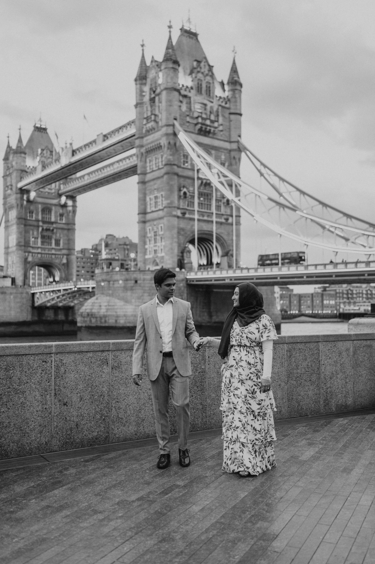 Black-and-white photograph of a couple holding hands on the Queen’s Walk with Tower Bridge behind during their London engagement photoshoot.