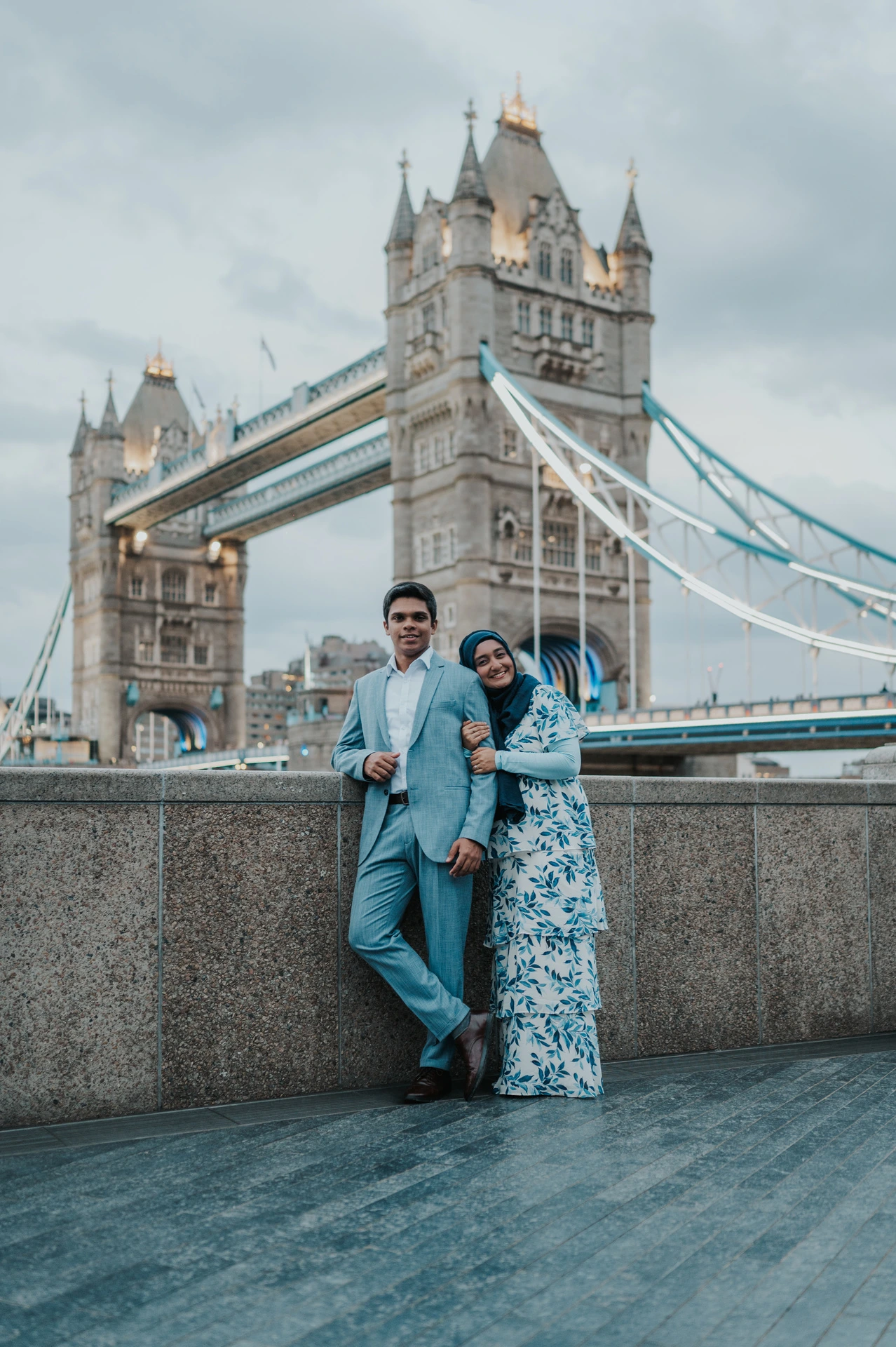 A smiling couple in blue outfits leaning together against the riverside wall near Tower Bridge during their London engagement photoshoot.