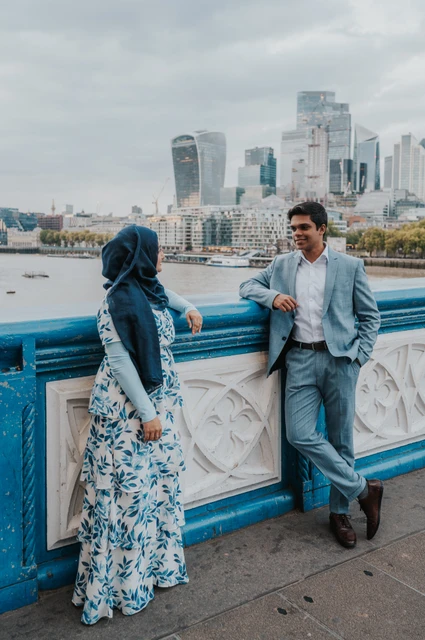 London engagement photoshoot: blue railings and the City skyline A couple facing each other beside Tower Bridge’s blue railing with skyscrapers across the Thames during their London engagement photoshoot.