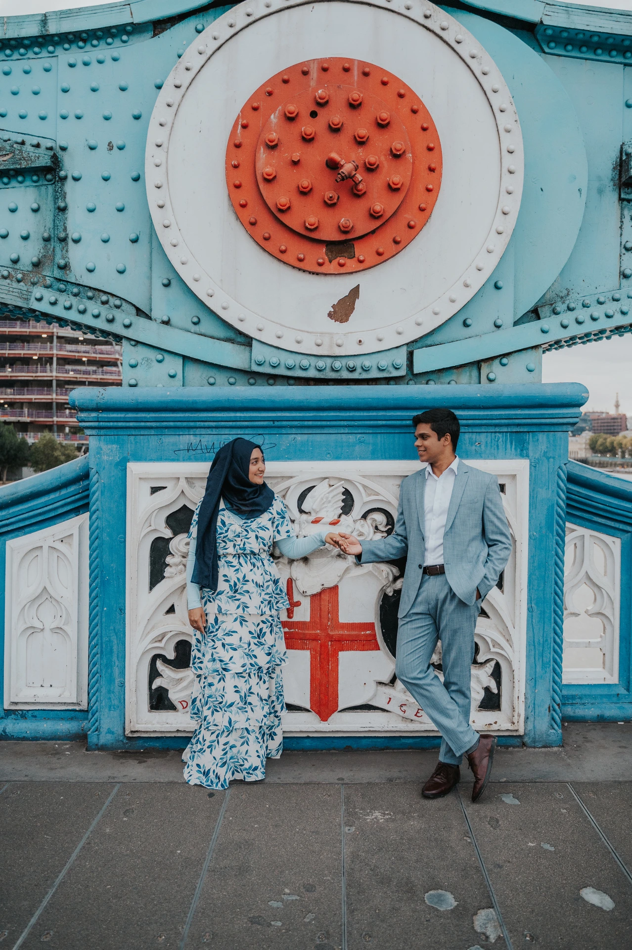 An engaged couple holding hands below Tower Bridge’s blue steelwork and large red riveted disc during their London engagement photoshoot.