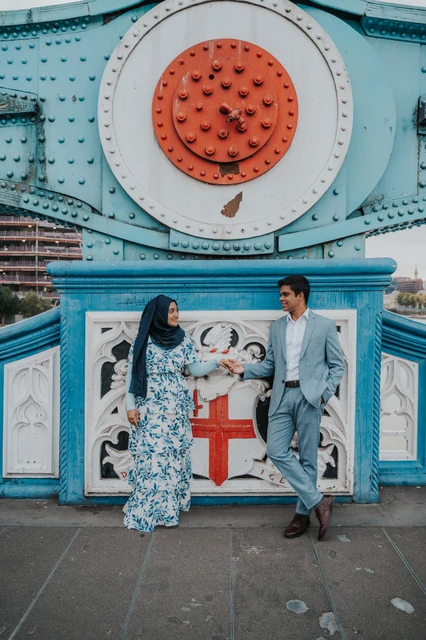 London engagement photoshoot: blue steelwork and a red bridge detail An engaged couple holding hands below Tower Bridge’s blue steelwork and large red riveted disc during their London engagement photoshoot.