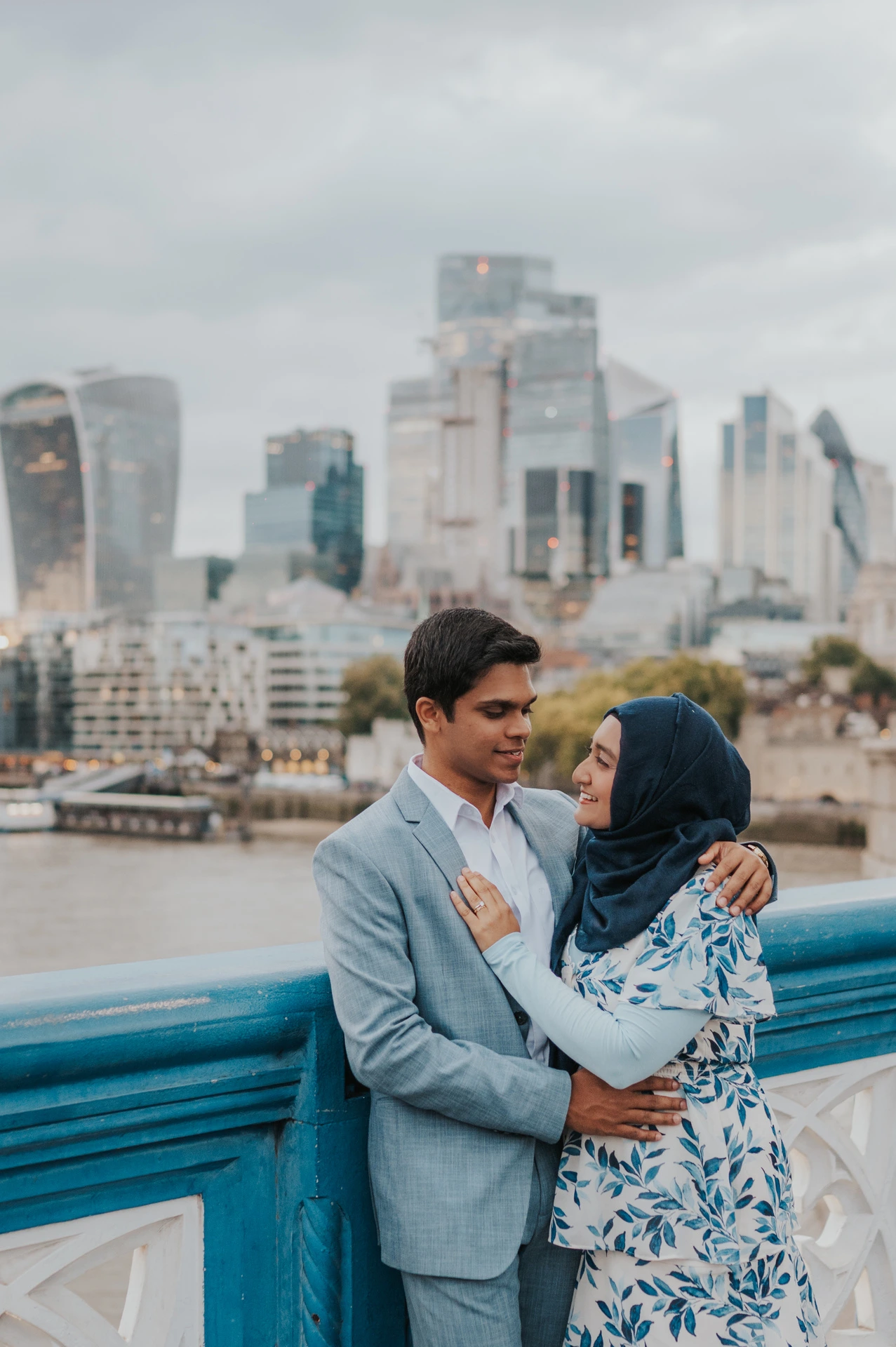 A couple smiling closely at each other above the blue railing with the City skyline beyond during their London engagement photoshoot.