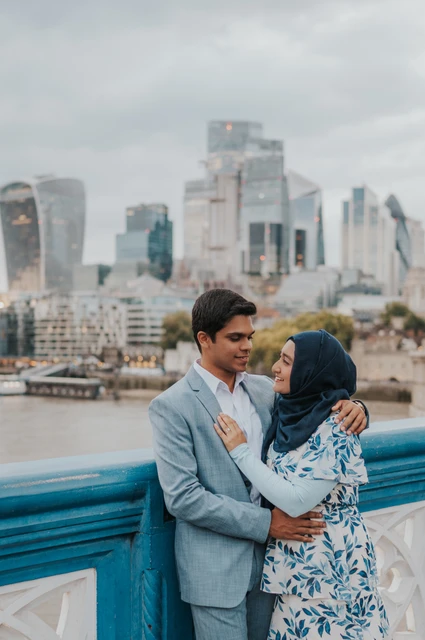 London engagement photoshoot: close smiles above the Thames A couple smiling closely at each other above the blue railing with the City skyline beyond during their London engagement photoshoot.