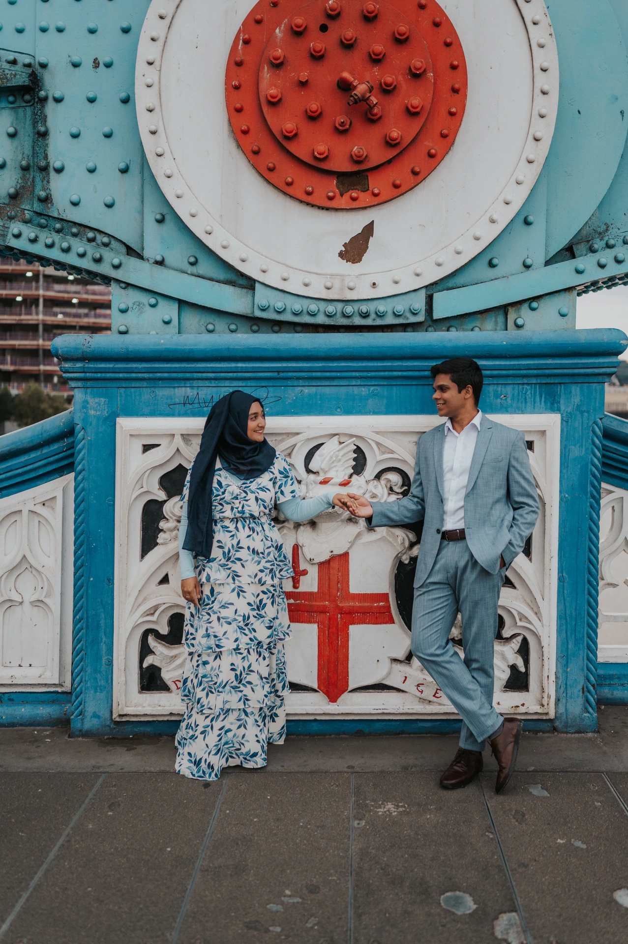 A couple holding hands beneath Tower Bridge’s blue beams and red circular detail during their London engagement photoshoot.