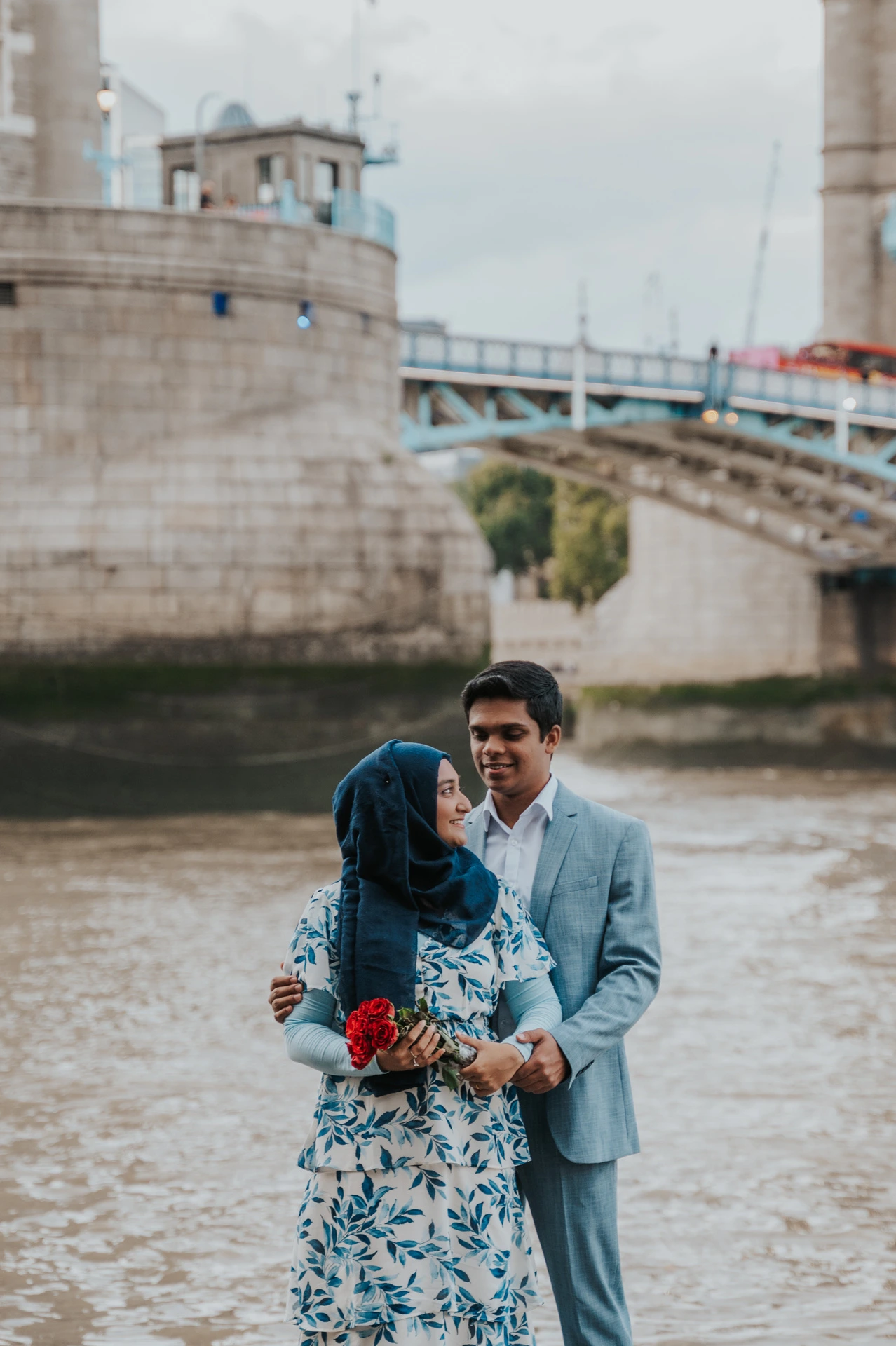A couple standing together with red roses beside the Thames and Tower Bridge during their London engagement photoshoot.