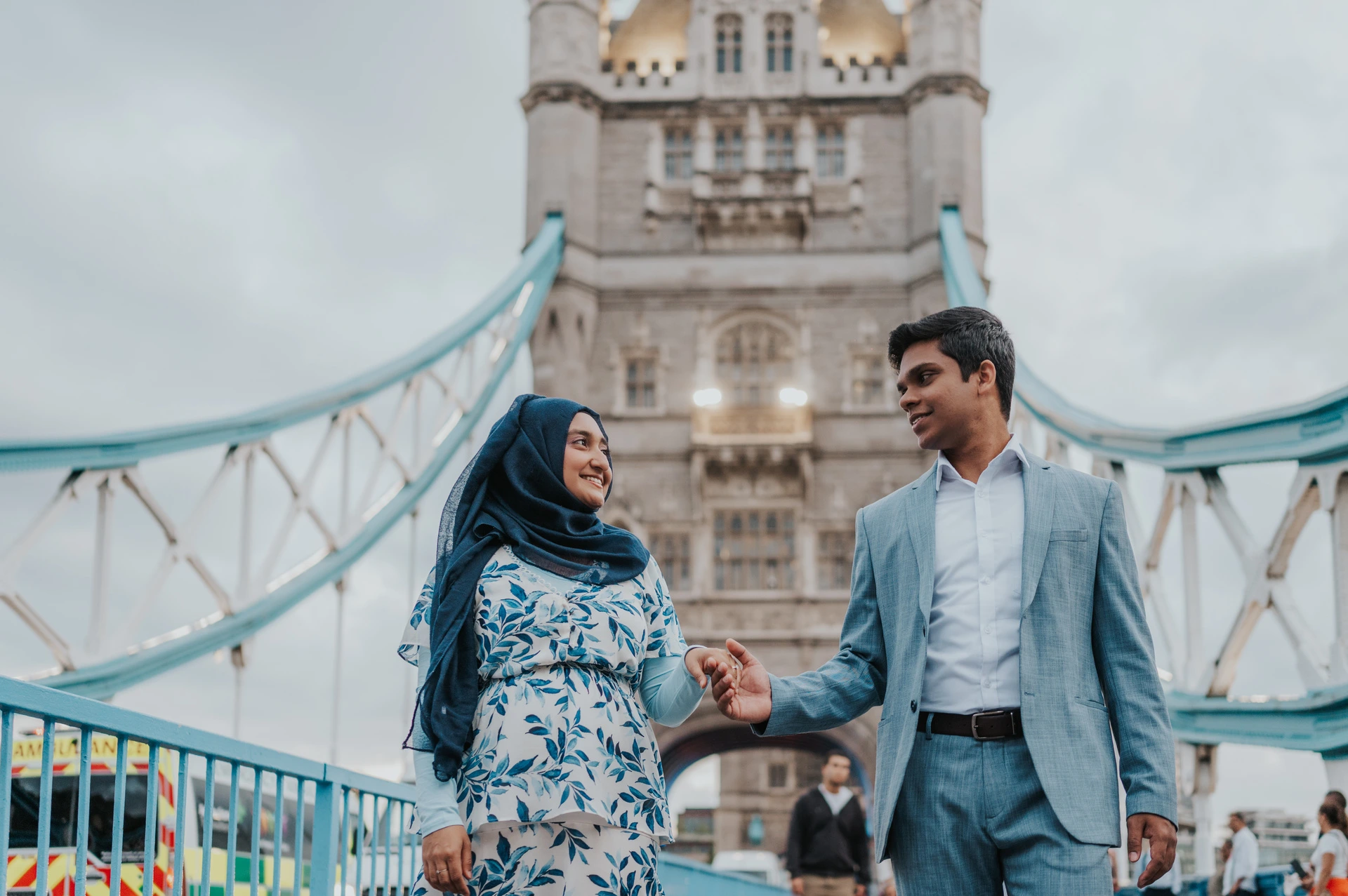 A smiling couple holding hands on Tower Bridge with blue suspension details and a stone tower behind them during their London engagement photoshoot.