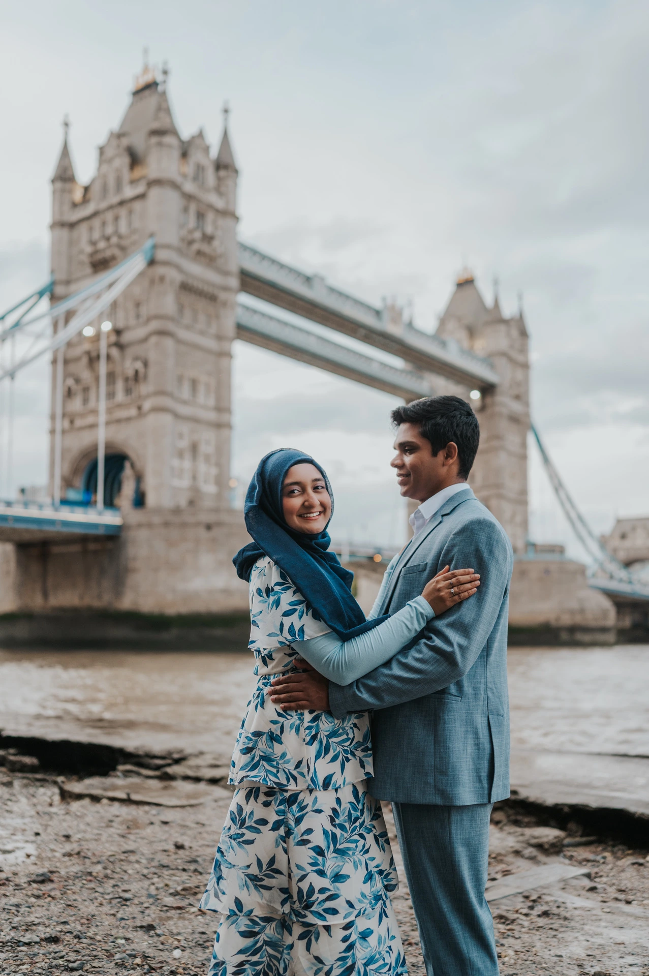 A smiling couple embracing in blue outfits with Tower Bridge behind them during their London engagement photoshoot.