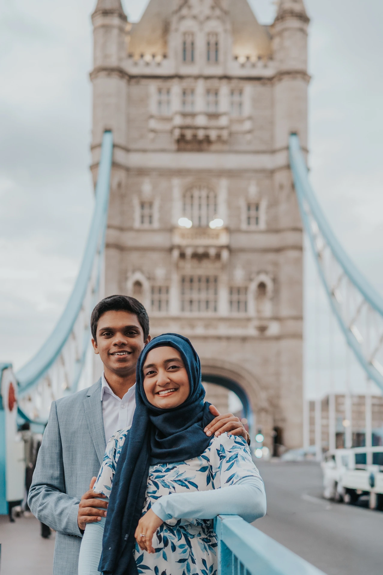 A smiling couple standing close on Tower Bridge with a stone tower softly blurred behind them during their London engagement photoshoot.