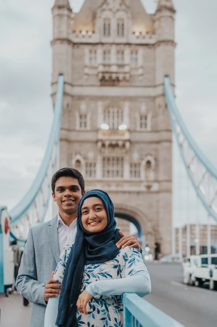 A smiling couple standing close on Tower Bridge with a stone tower softly blurred behind them during their London engagement photoshoot.