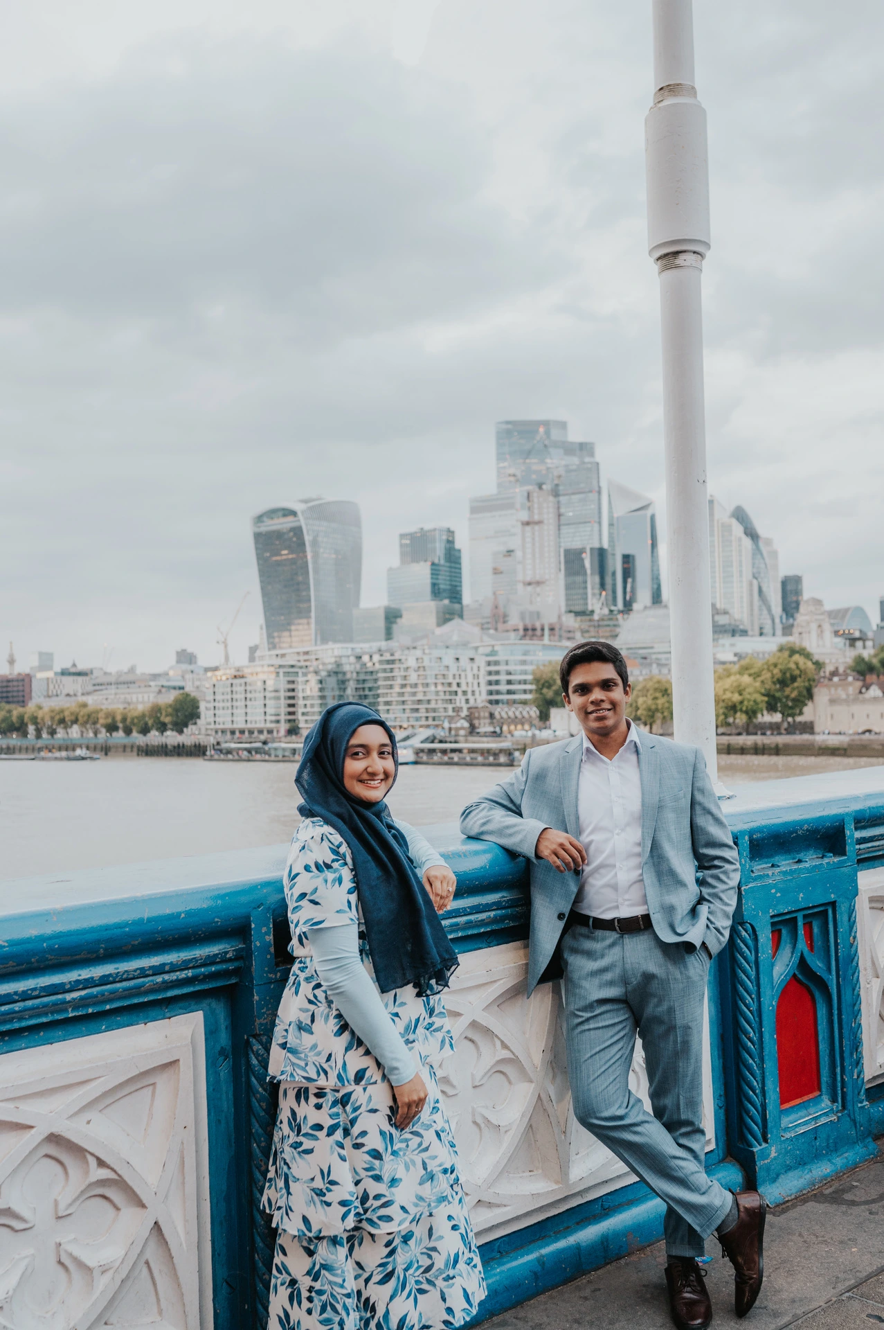 A smiling couple beside Tower Bridge’s blue and white railing with the City skyline across the Thames during their London engagement photoshoot.