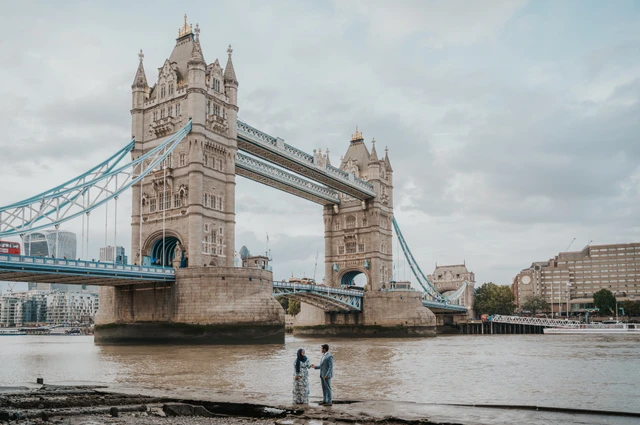 Wide view of an engaged couple beside the Thames with both towers of Tower Bridge above them during their London engagement photoshoot.