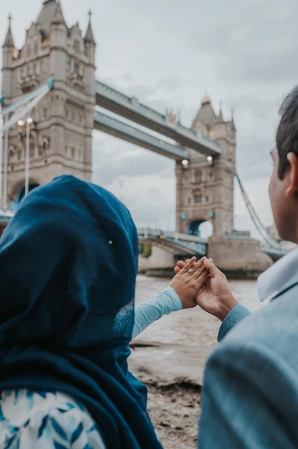 London engagement photoshoot: their engagement ring and the bridge towers A couple seen from behind holding up their joined hands towards Tower Bridge during their London engagement photoshoot.