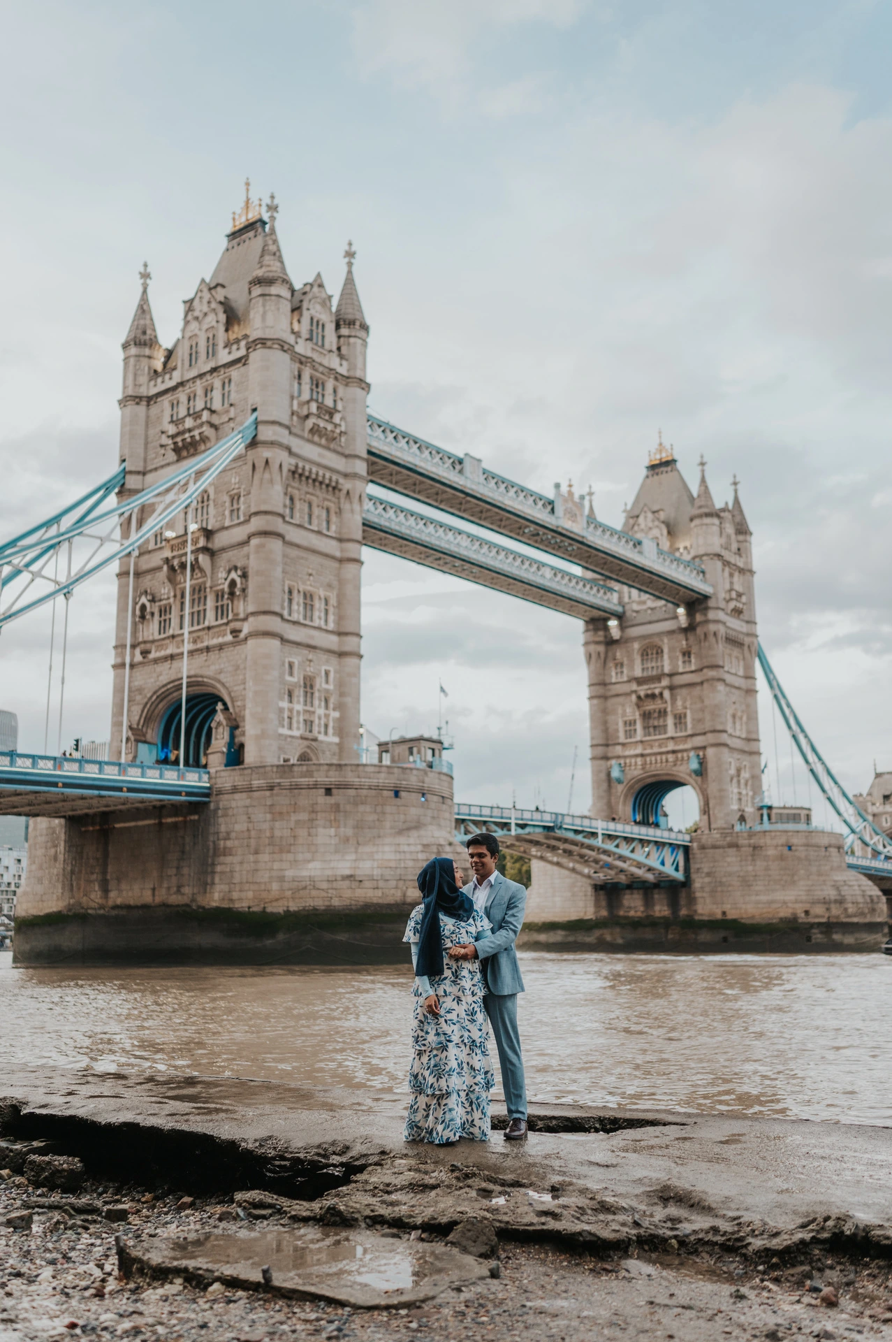 A couple standing close with Tower Bridge rising above the Thames behind them during their London engagement photoshoot.