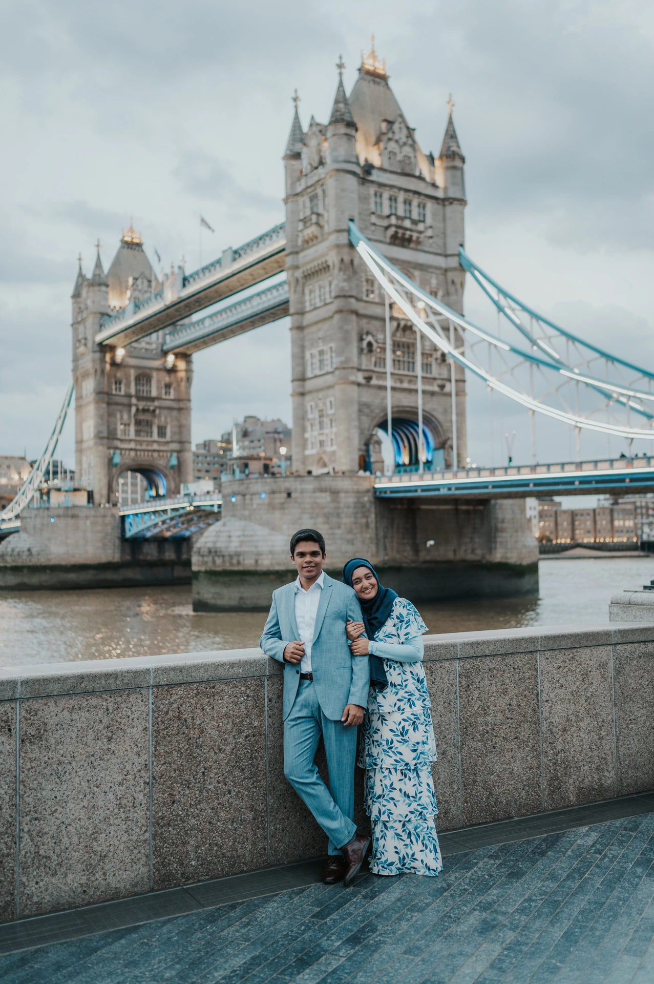 A couple leaning against the riverside wall with both towers of Tower Bridge in view during their London engagement photoshoot.