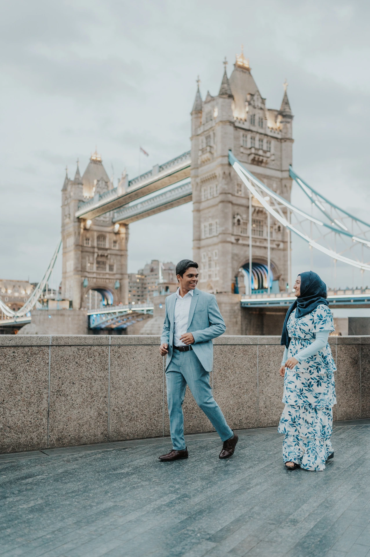 A couple walking together beside the Queen’s Walk riverside wall below Tower Bridge during their London engagement photoshoot.