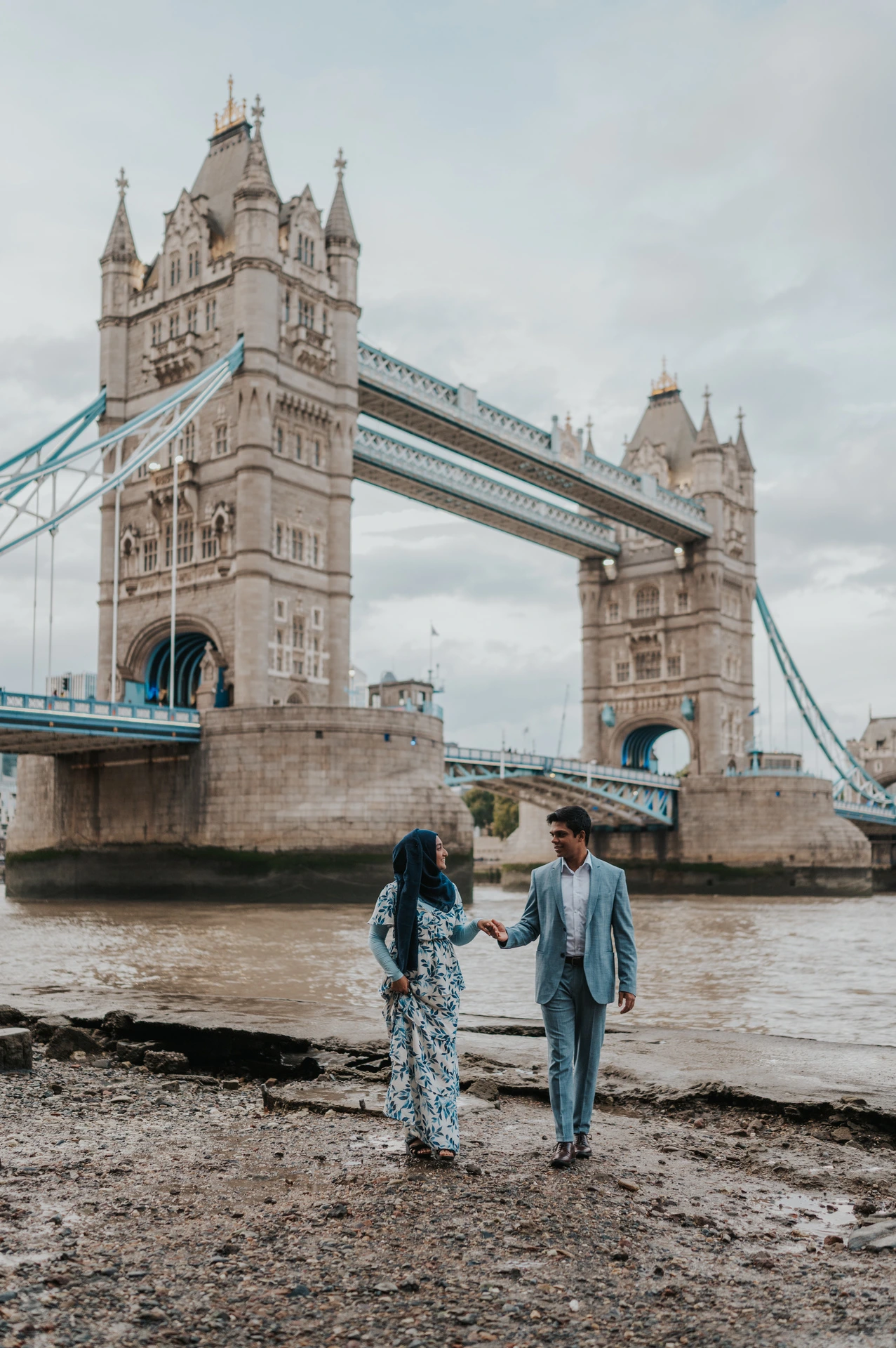 A couple holding hands as they walk across the Thames foreshore below Tower Bridge during their London engagement photoshoot.