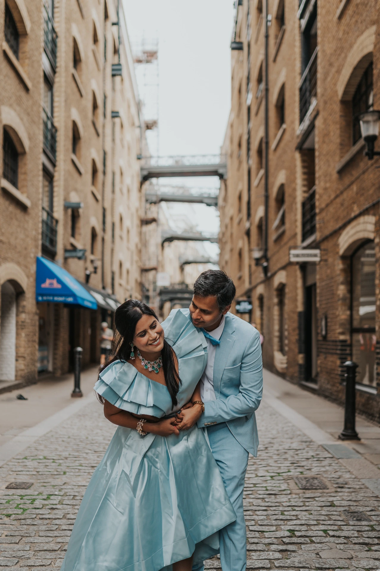 A couple in pale blue outfits embracing in a cobbled lane between brick warehouse buildings during their London family photoshoot.