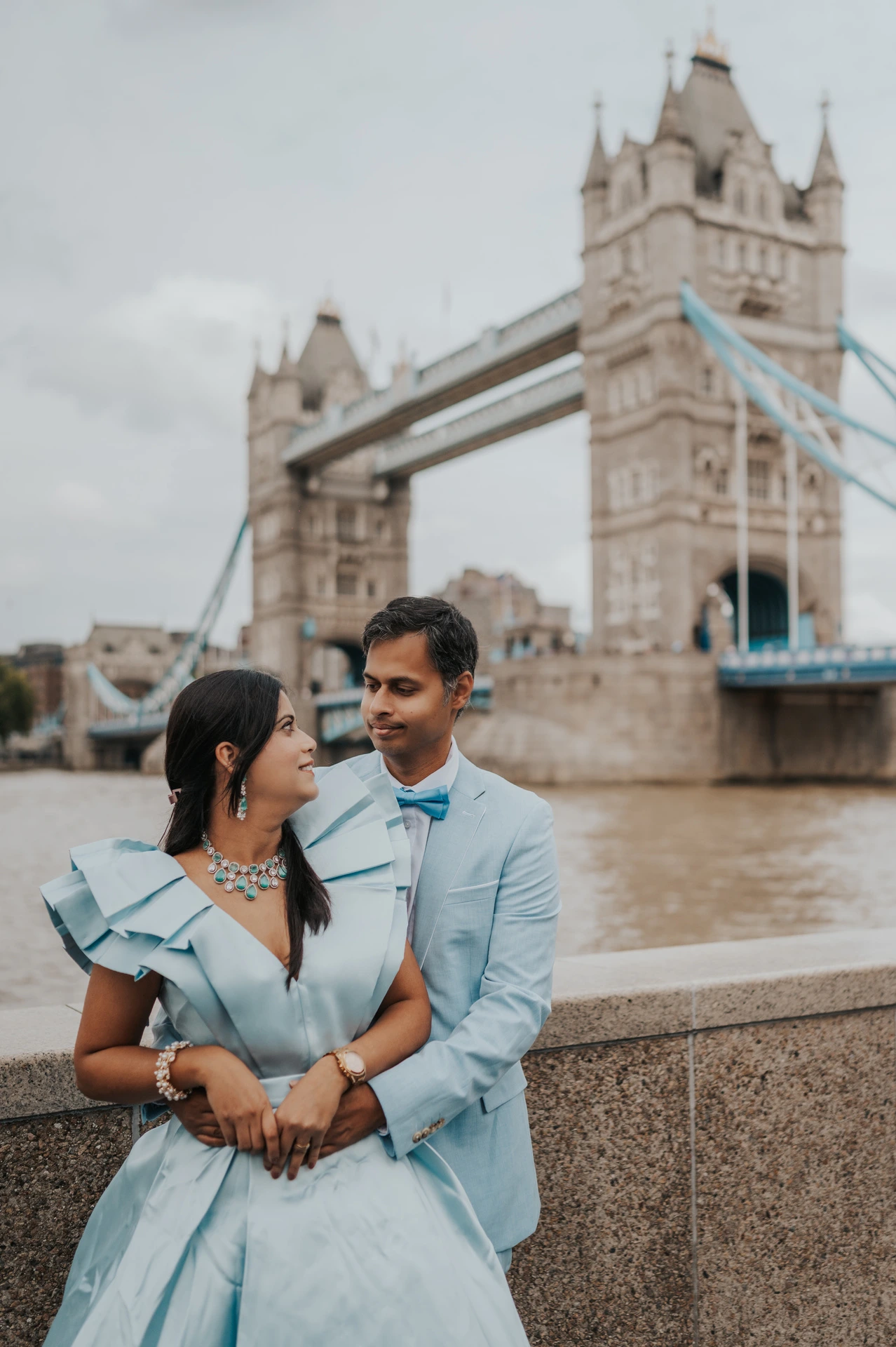 A couple in pale blue outfits standing close beside the Thames and looking towards each other during their London family photoshoot.