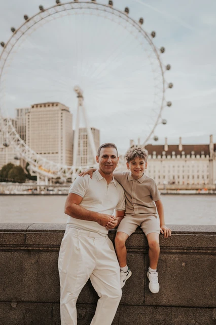 London family photoshoot: a close family portrait with the London Eye A father holding his child beside a riverside wall with the London Eye behind them during their London family photoshoot.