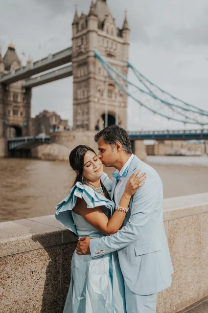 London family photoshoot: a close portrait beside Tower Bridge A couple leaning close beside a stone riverside wall with Tower Bridge behind them during their London family photoshoot.