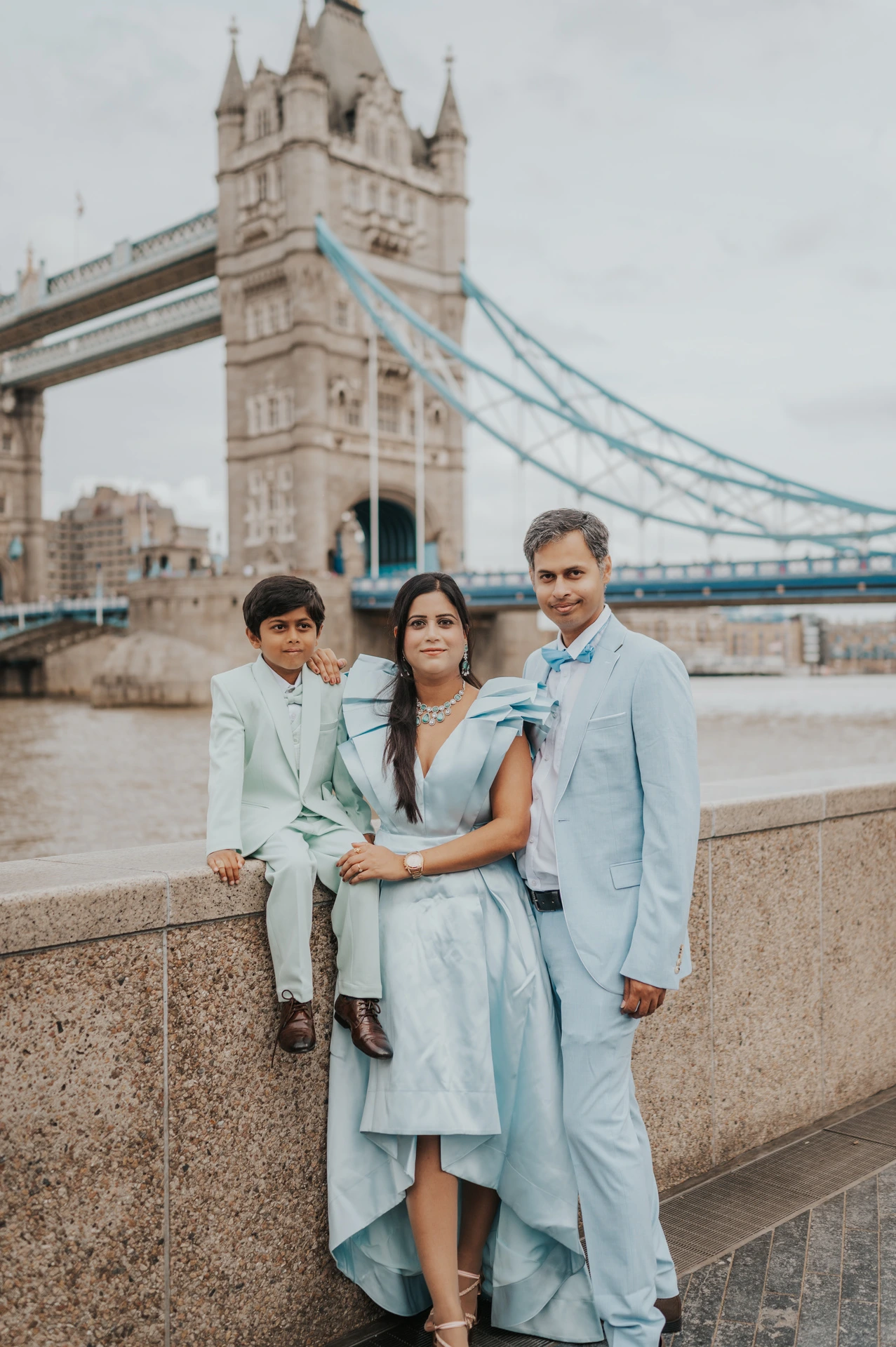 A closer family portrait beside the Thames with Tower Bridge rising behind the three of them during their London family photoshoot.