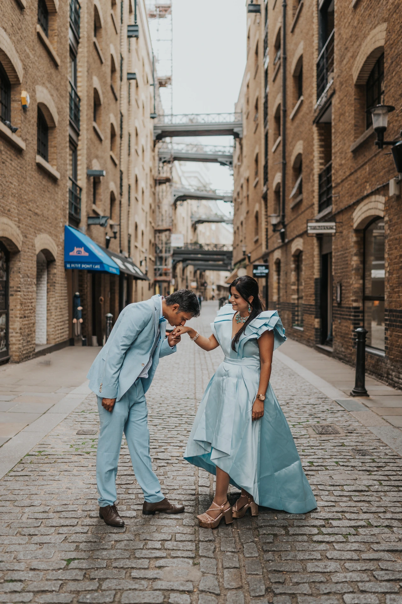 A man bending towards his partner’s hand in a cobbled lane with brick buildings and overhead walkways during their London family photoshoot.