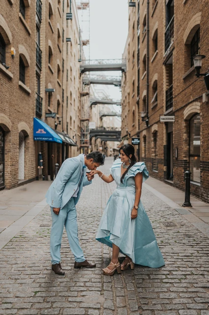 London family photoshoot: a couple portrait beneath the overhead walkways A man bending towards his partner’s hand in a cobbled lane with brick buildings and overhead walkways during their London family photoshoot.