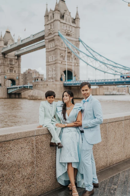 London family photoshoot: a family in blue and mint beside Tower Bridge A child sitting on a stone riverside wall beside two adults in pale blue outfits with Tower Bridge behind during their London family photoshoot.
