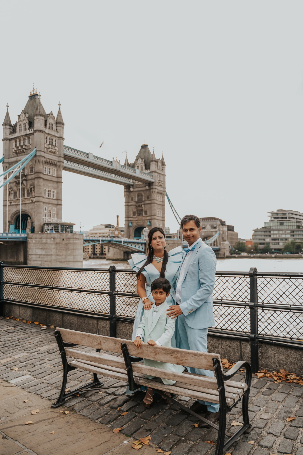 A family of three gathered beside a wooden bench with Tower Bridge across the Thames behind them during their London family photoshoot.
