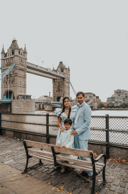 London family photoshoot: a family moment beside the riverside bench A family of three gathered beside a wooden bench with Tower Bridge across the Thames behind them during their London family photoshoot.
