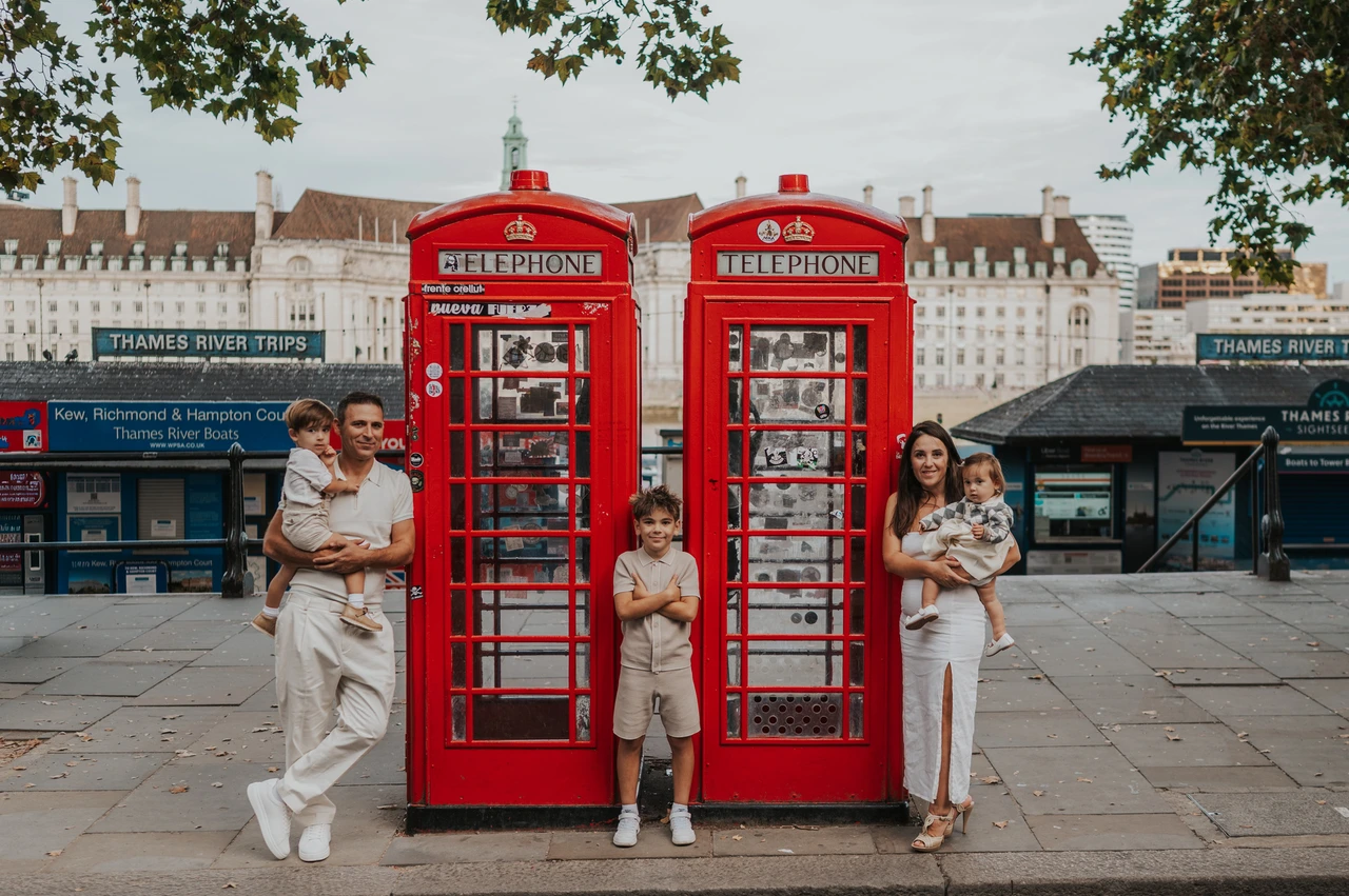 London family photoshoot: a family portrait at the red telephone boxes Parents holding two children beside red telephone boxes while another child stands between the boxes during their London family photoshoot.