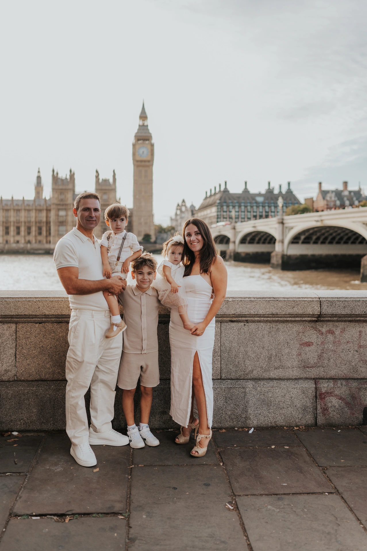 A family of five standing beside the Thames with Big Ben and Westminster Bridge behind them during their London family photoshoot.