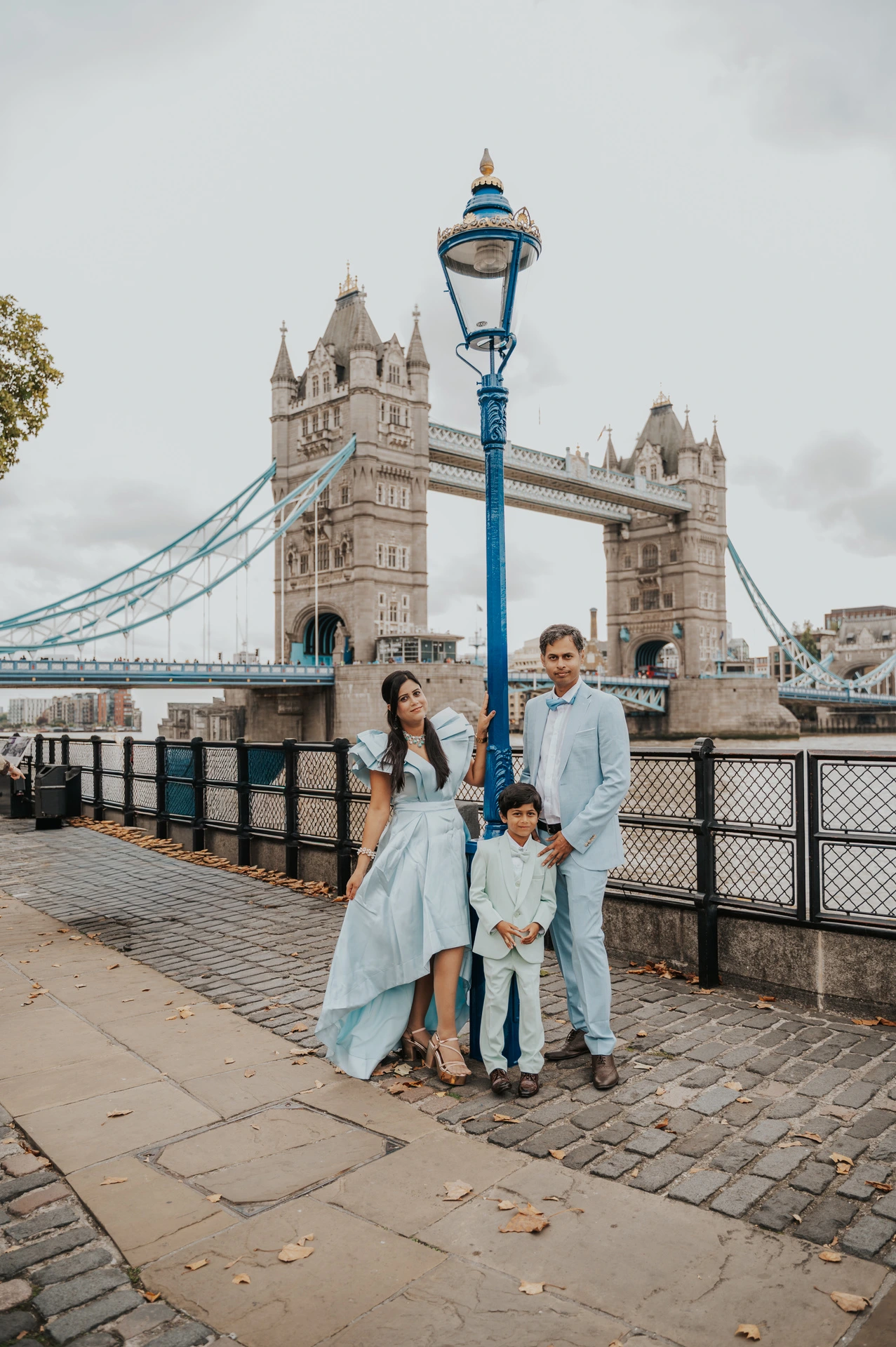A family of three standing beside dark riverside railings and a lamp post with Tower Bridge beyond during their London family photoshoot.