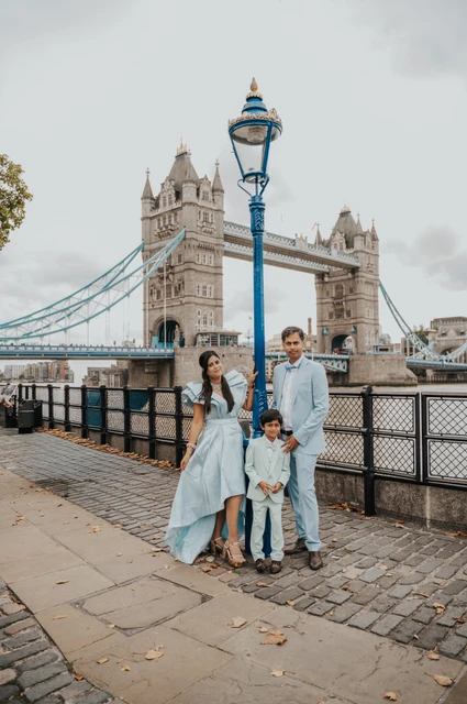 London family photoshoot: a family portrait beside the riverside lamp post A family of three standing beside dark riverside railings and a lamp post with Tower Bridge beyond during their London family photoshoot.