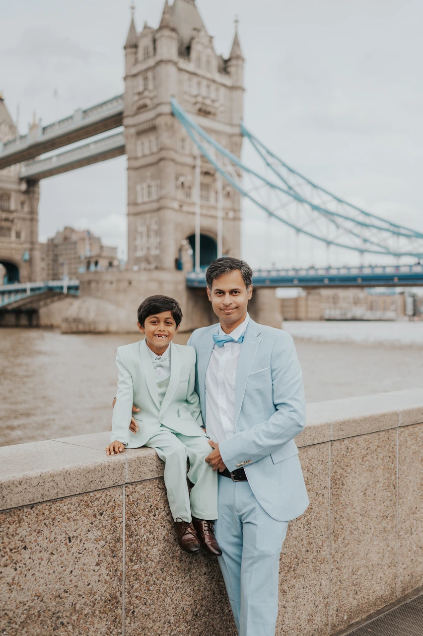 London family photoshoot: a father-and-child portrait by Tower Bridge A man in a pale blue suit standing beside a boy sitting on a riverside wall below Tower Bridge during their London family photoshoot.