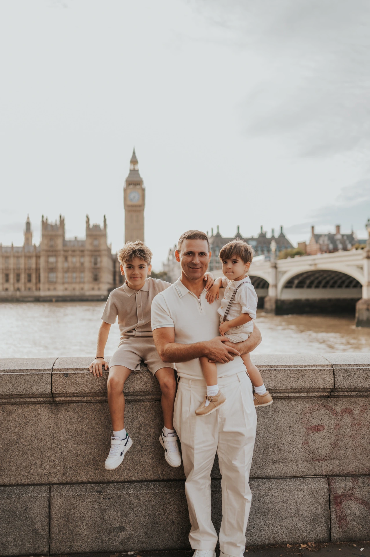 A father sitting on a riverside wall with two children and Big Ben across the Thames during their London family photoshoot.