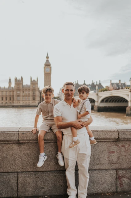 A father sitting on a riverside wall with two children and Big Ben across the Thames during their London family photoshoot.