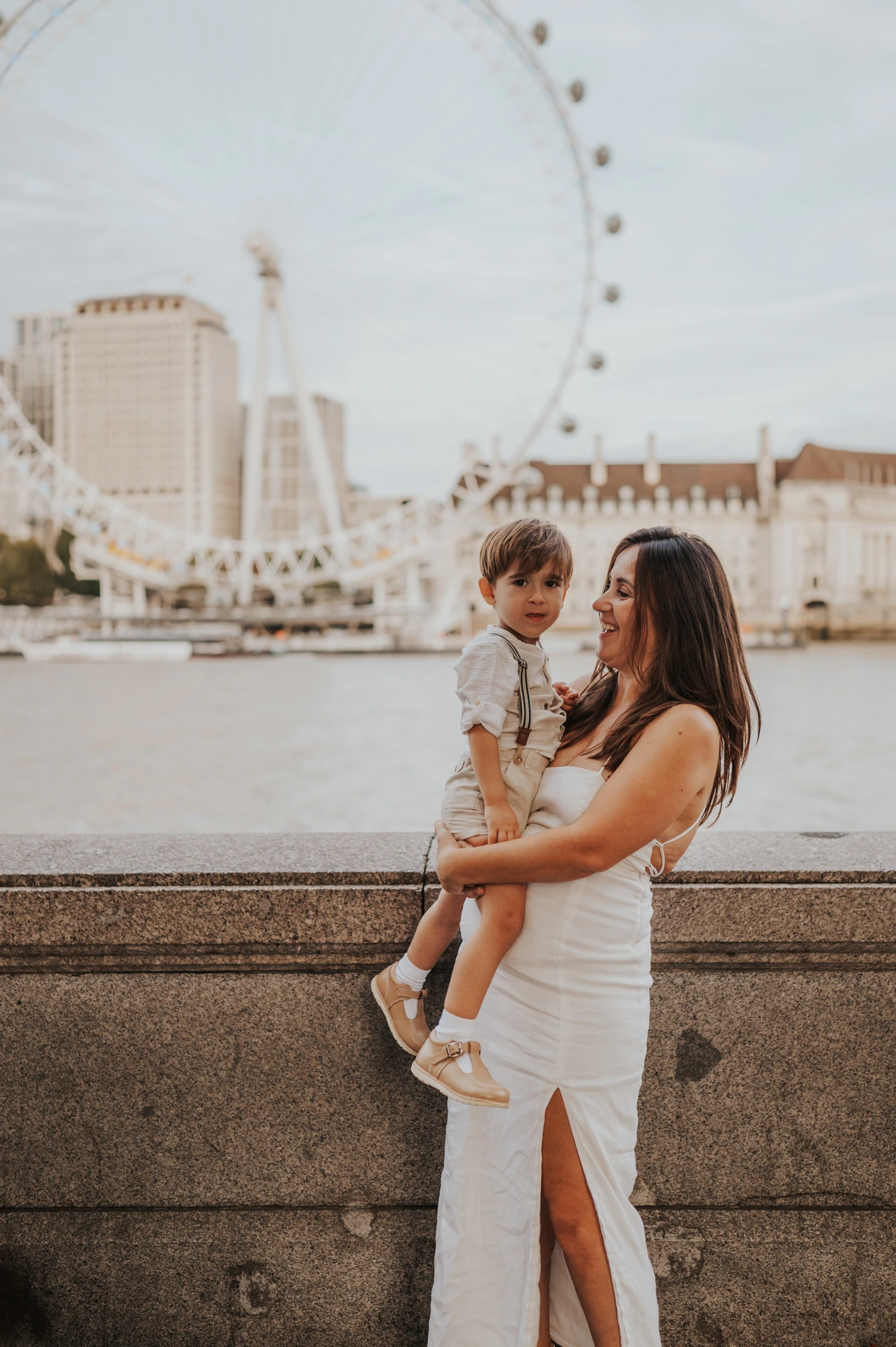 A mother looking at the child in her arms beside the river with the London Eye behind during their London family photoshoot.