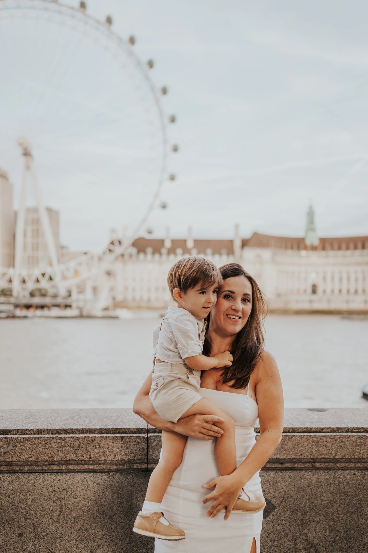 A mother holding her child beside the Thames with the London Eye softly blurred behind them during their London family photoshoot.