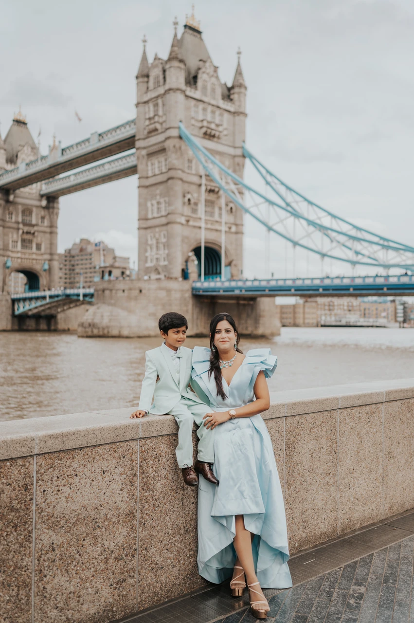 London family photoshoot: a mother-and-child portrait beside the river A woman in a pale blue dress standing beside a boy sitting on the riverside wall with Tower Bridge behind during their London family photoshoot.