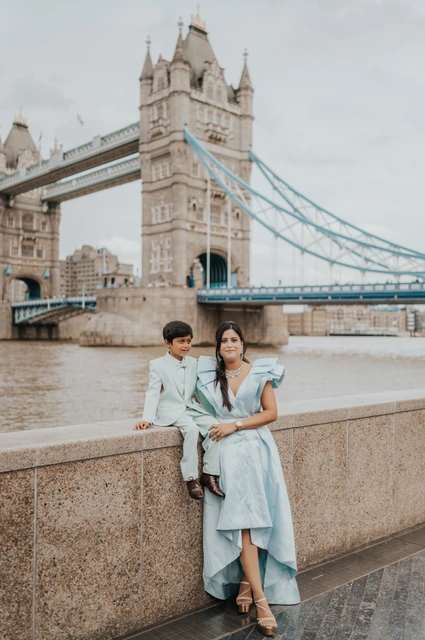 A woman in a pale blue dress standing beside a boy sitting on the riverside wall with Tower Bridge behind during their London family photoshoot.