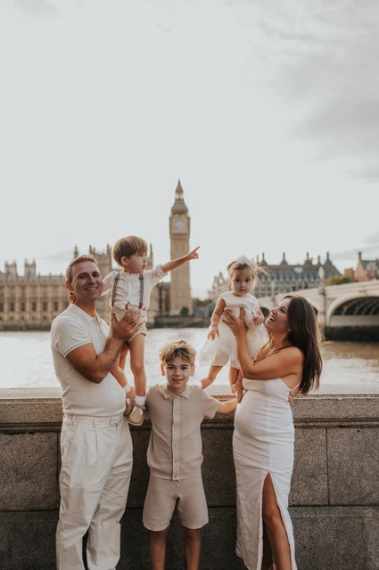 London family photoshoot: a playful family moment beside Big Ben Parents and three children sharing a playful moment beside the Thames with Big Ben in view during their London family photoshoot.