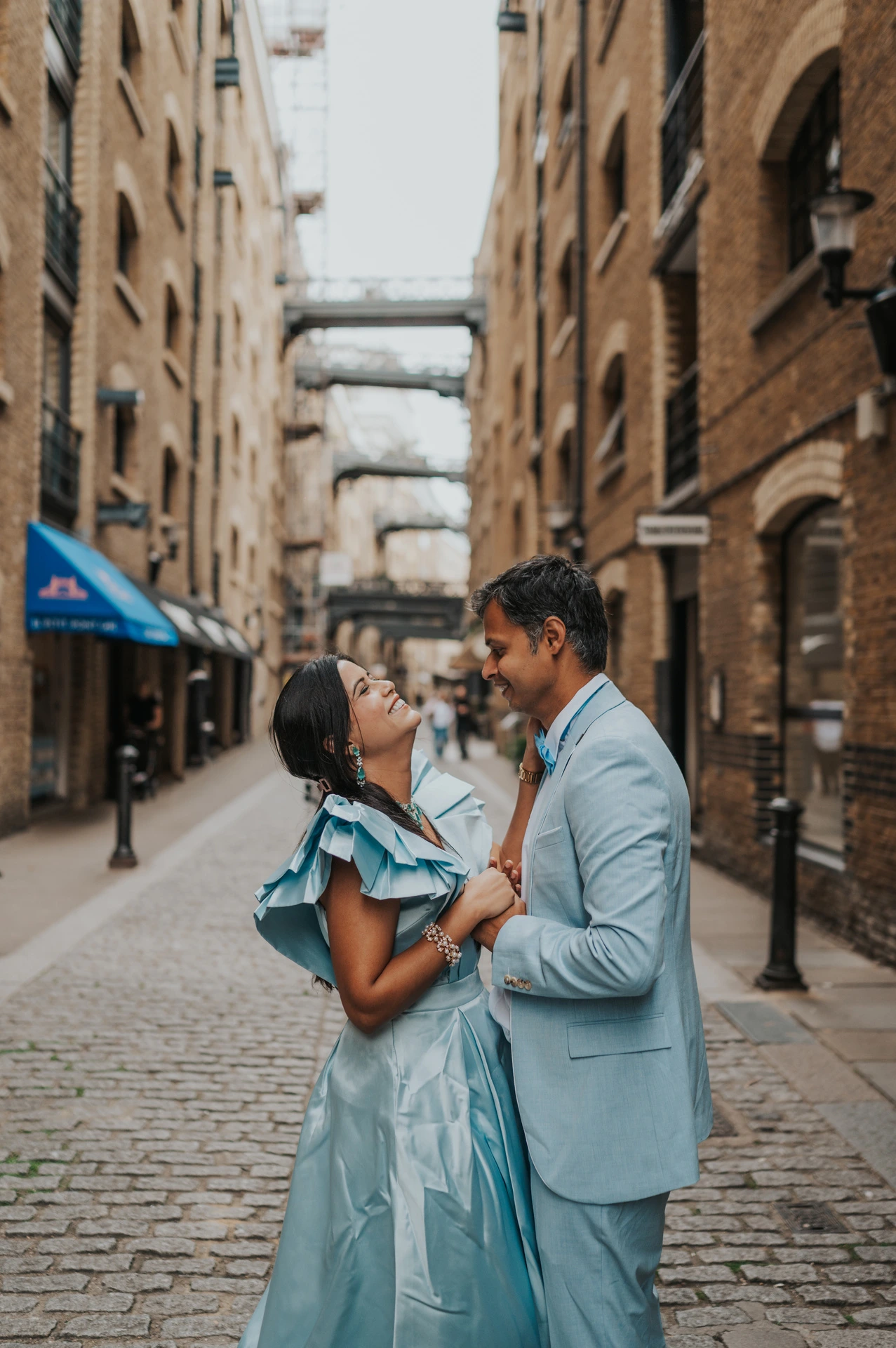A couple looking at each other in a cobbled lane with overhead walkways and brick walls during their London family photoshoot.