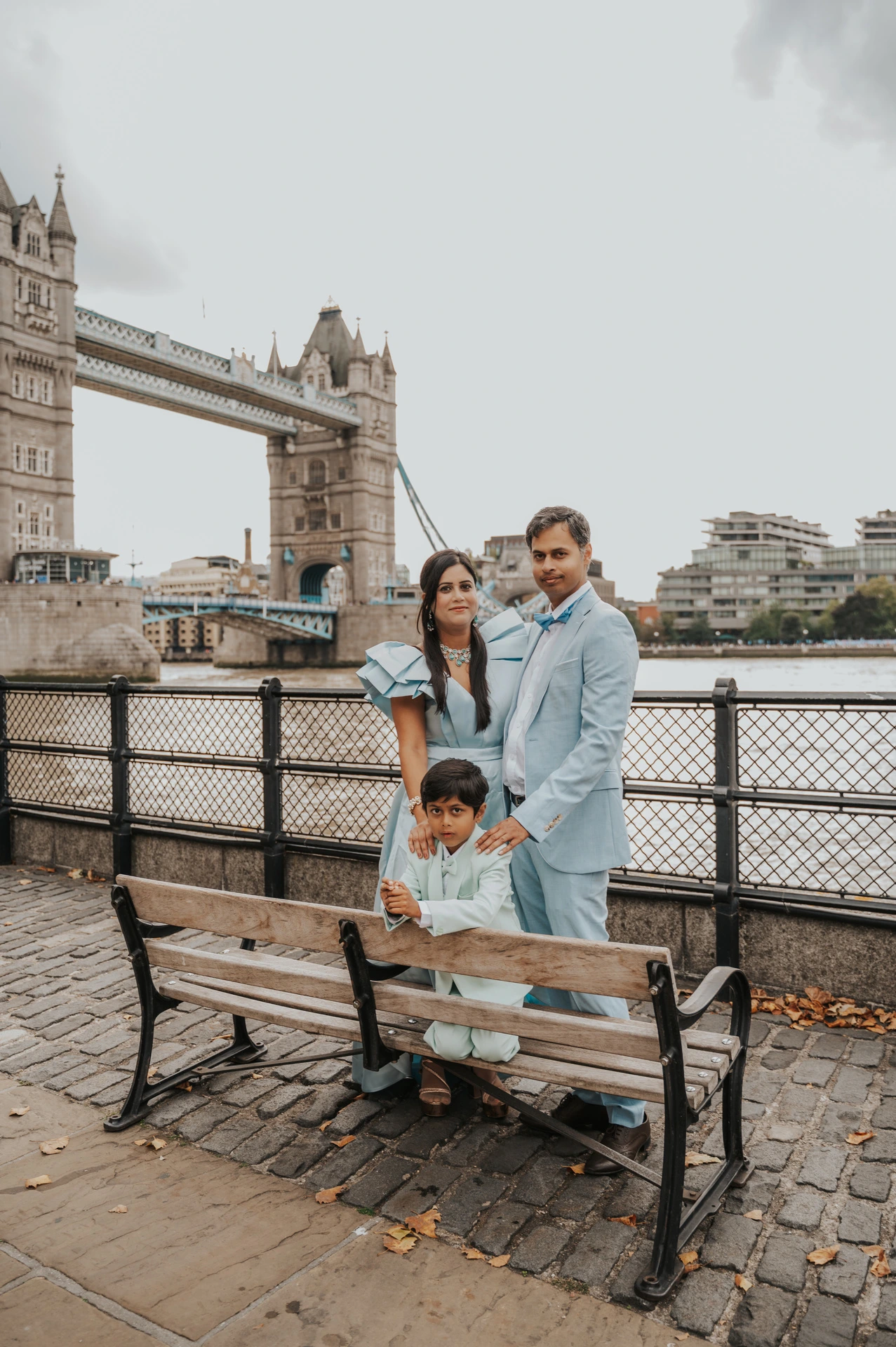 A couple looking towards a child seated on a wooden bench with Tower Bridge in the background during their London family photoshoot.