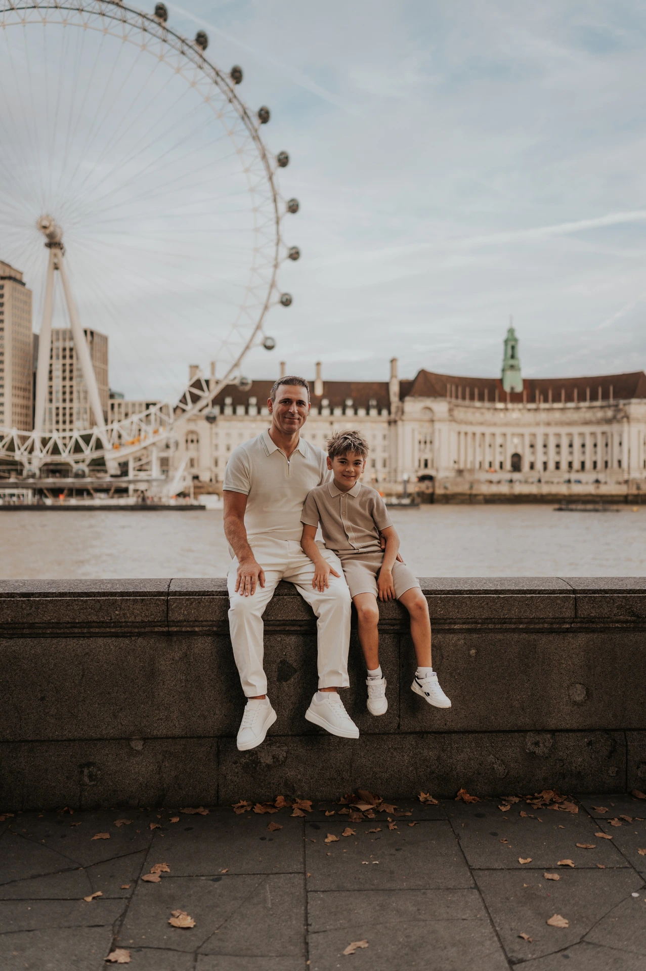 A father and child sitting together on the riverside wall beneath the London Eye during their London family photoshoot.