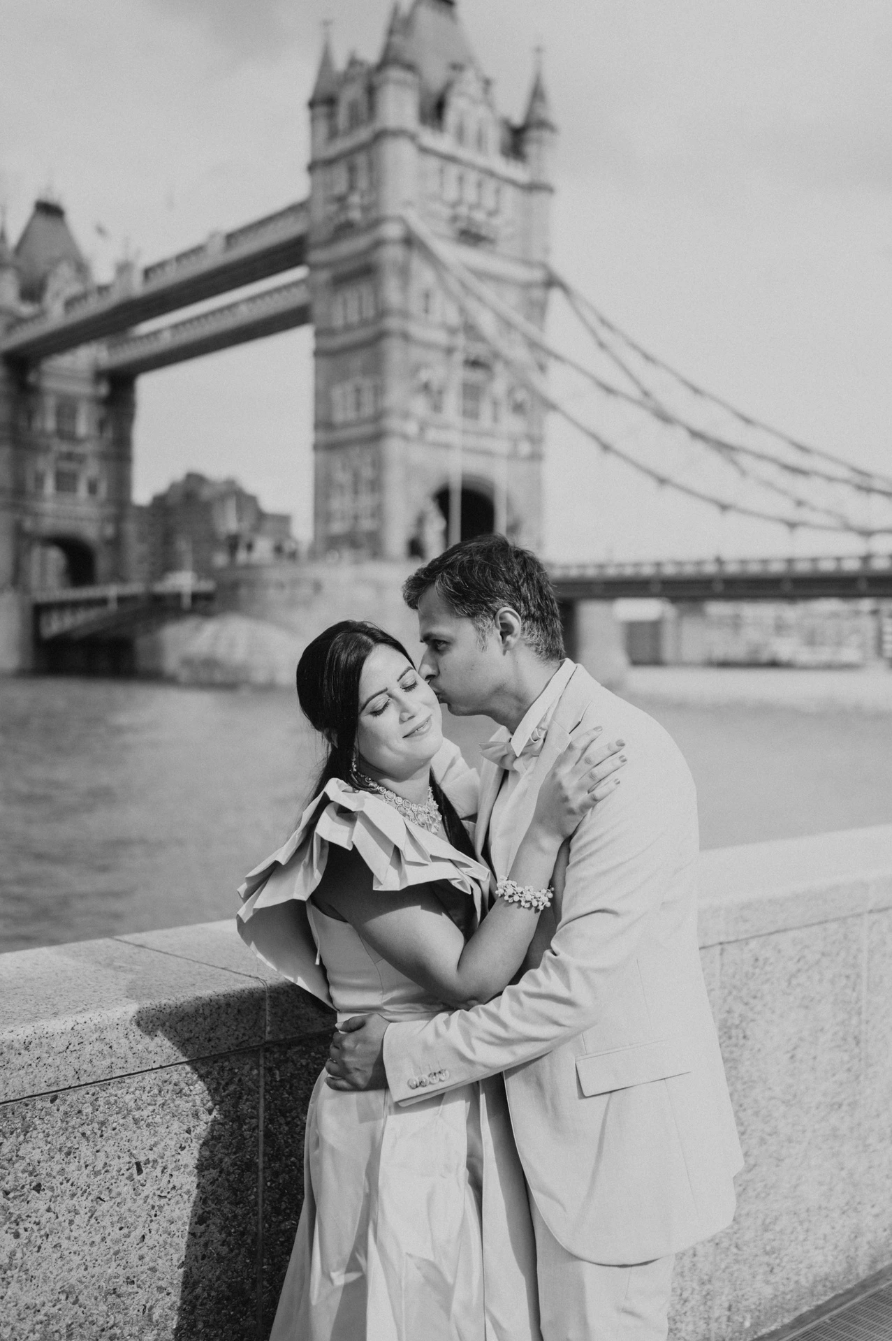 Black-and-white portrait of a couple leaning close beside the riverside wall and Tower Bridge during their London family photoshoot.