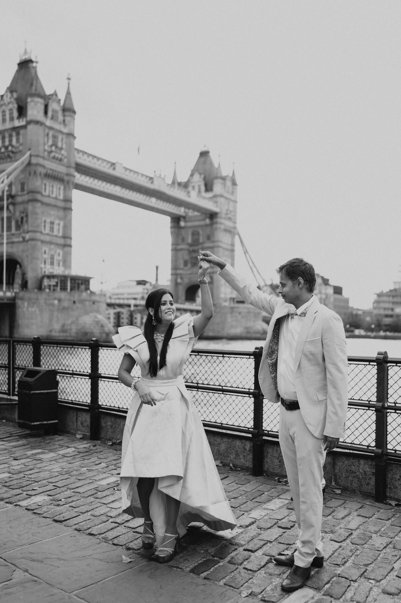 Black-and-white portrait of a couple holding hands beside a wooden bench and dark riverside railings during their London family photoshoot.