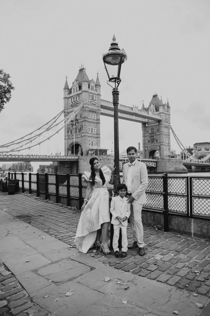 Black-and-white portrait of a family beside a riverside lamp post and railings near Tower Bridge during their London family photoshoot.