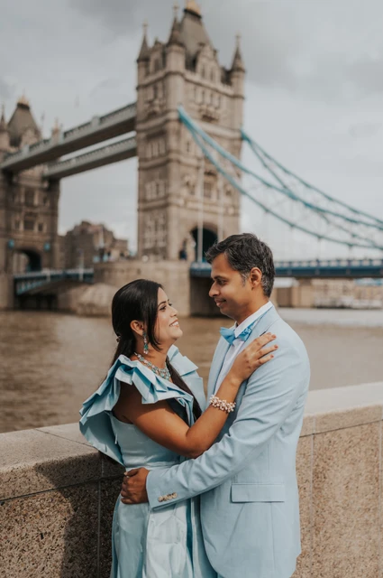 London family photoshoot: blue outfits against the river and bridge A couple in pale blue outfits smiling at each other beside the Thames with Tower Bridge behind during their London family photoshoot.
